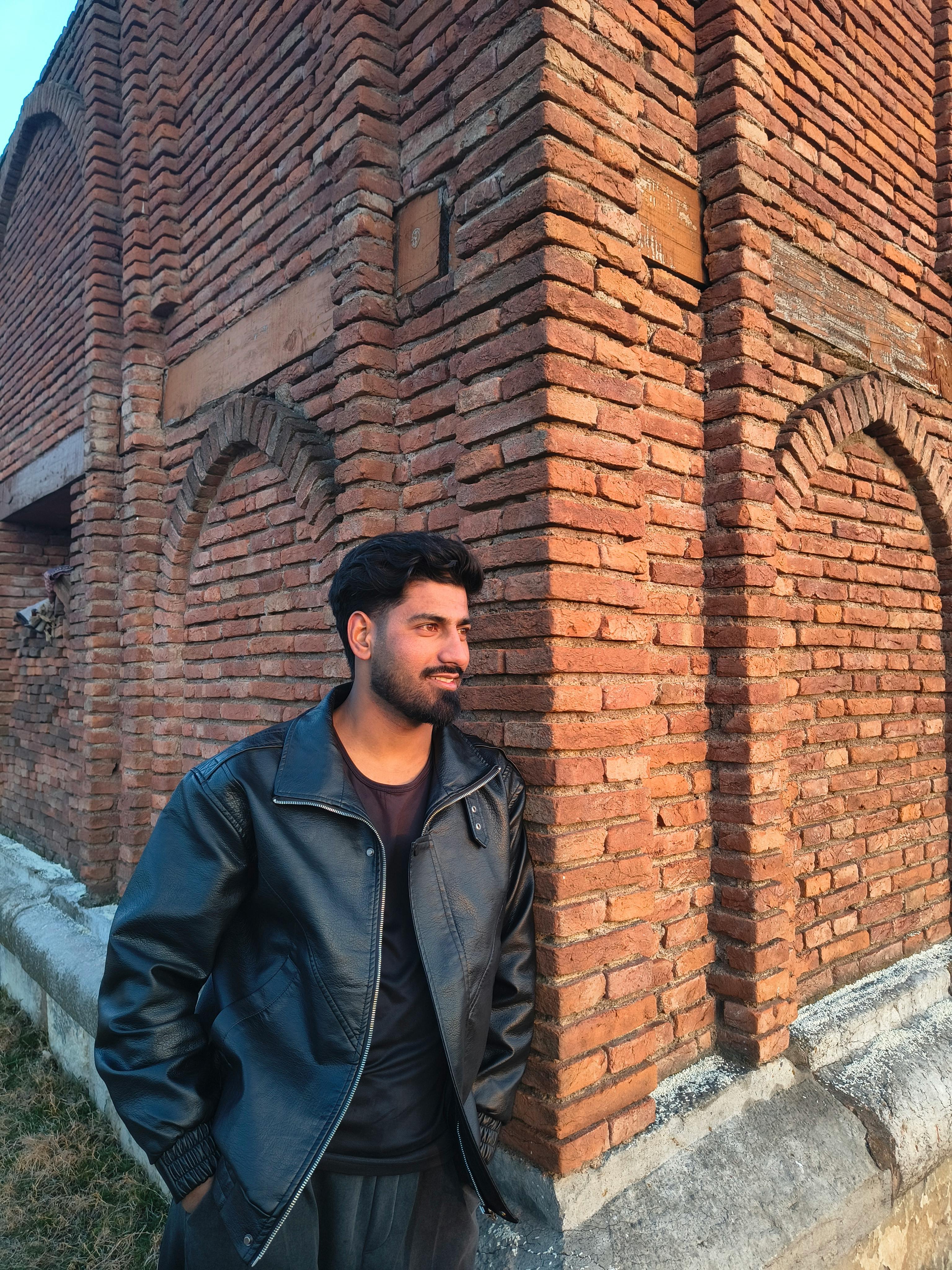 Man in black leather jacket beside a historic brick wall in daylight.