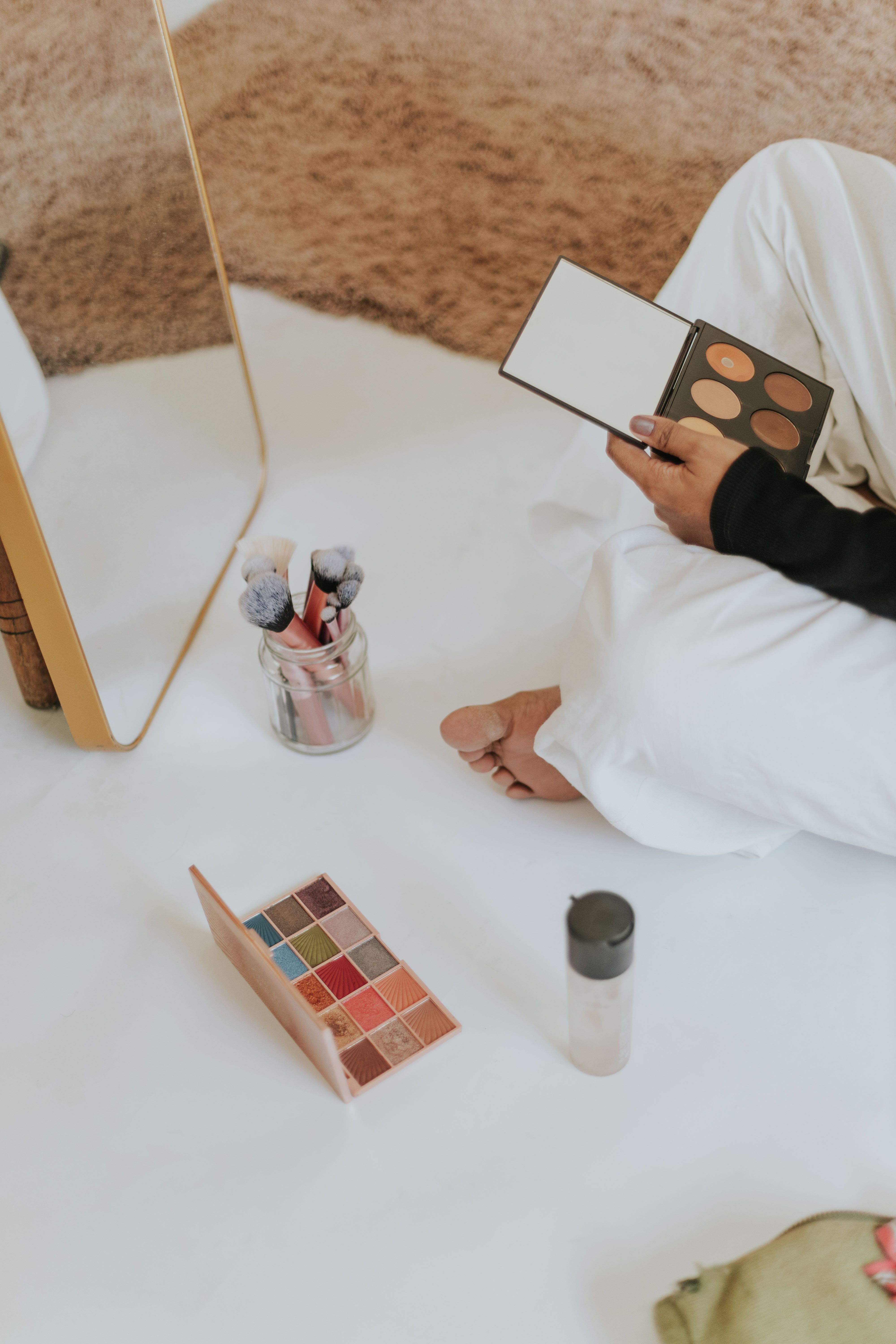 Woman sitting on floor applying makeup using various cosmetics, including palette and brushes.