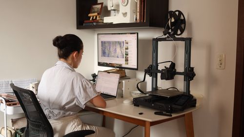 Free A woman working on a laptop with a 3D printer in a modern home office setup. Stock Photo