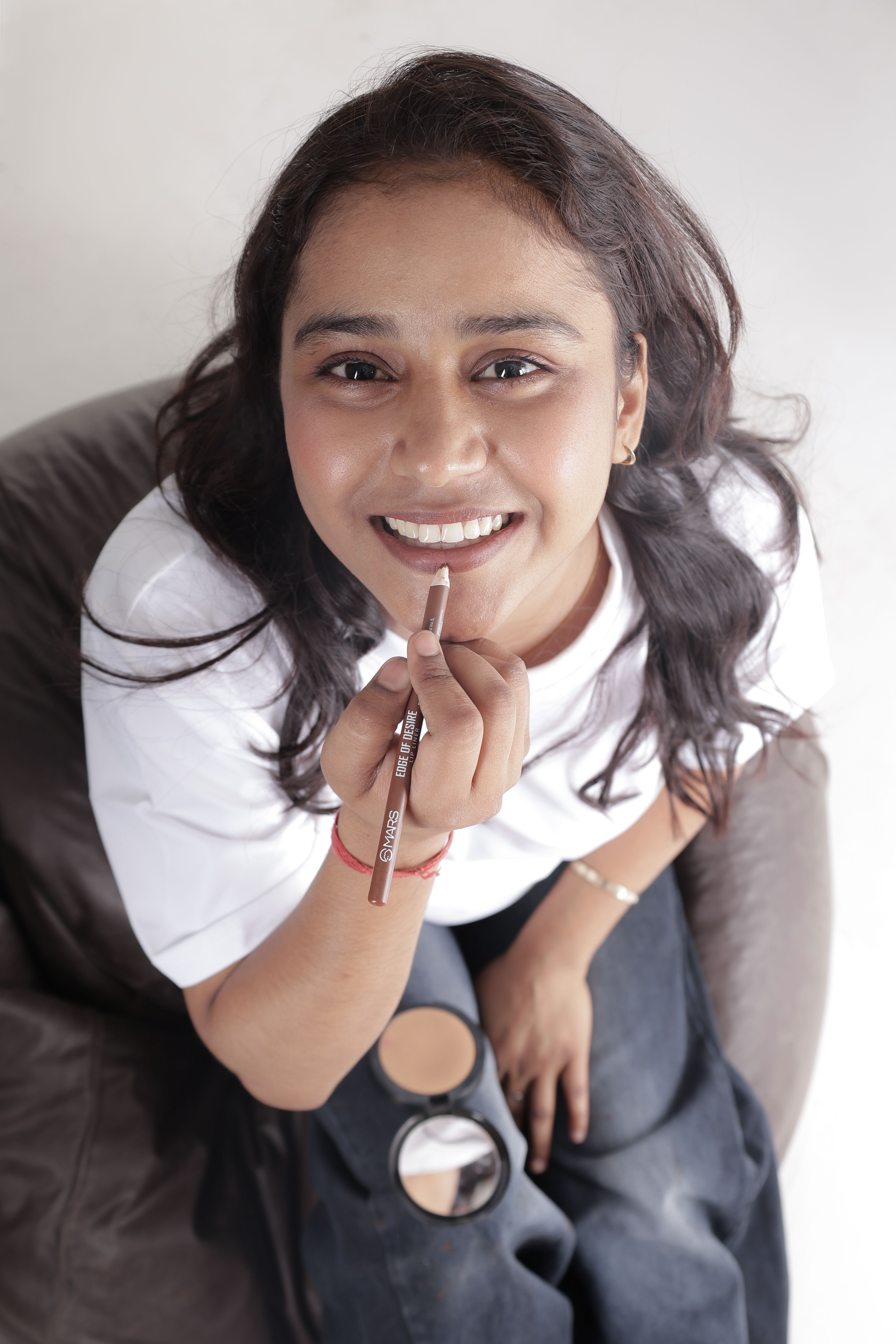 Cheerful woman applying makeup in casual attire, looking up with a smile.