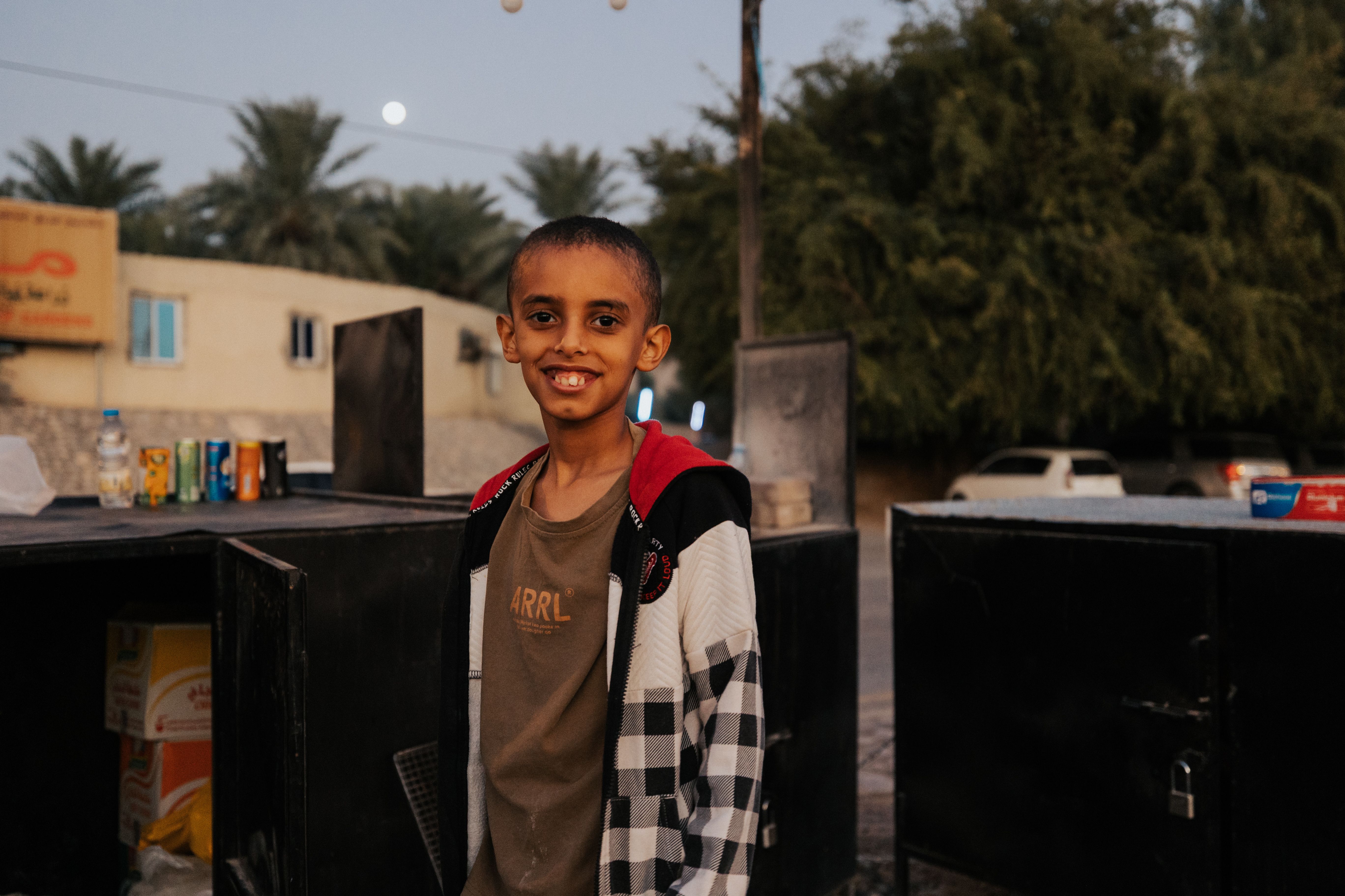 A smiling young boy stands at a street vendor stand in Nizwa, Oman during a warm day.