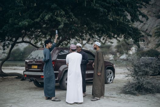 A group of Omani men socializing under a tree by a parked vehicle in Nizwa, Oman.