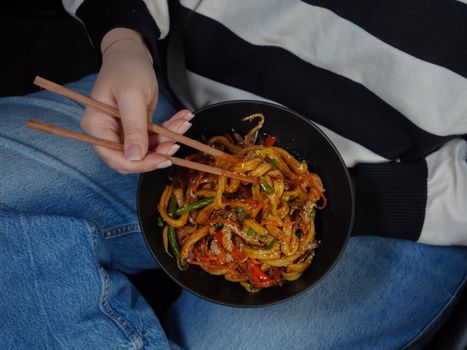 A person holding a bowl of stir-fried noodles with chopsticks, showcasing colorful vegetables.