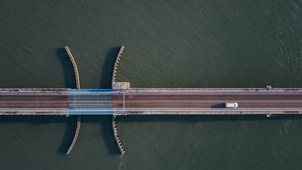 一张自上而下的航拍照片中，一辆白色汽车正行驶在跨越水面的桥梁上