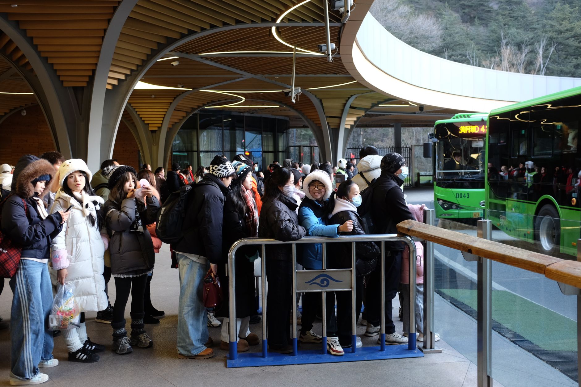 A large group of people in winter clothing wait for a bus under a modern shelter.