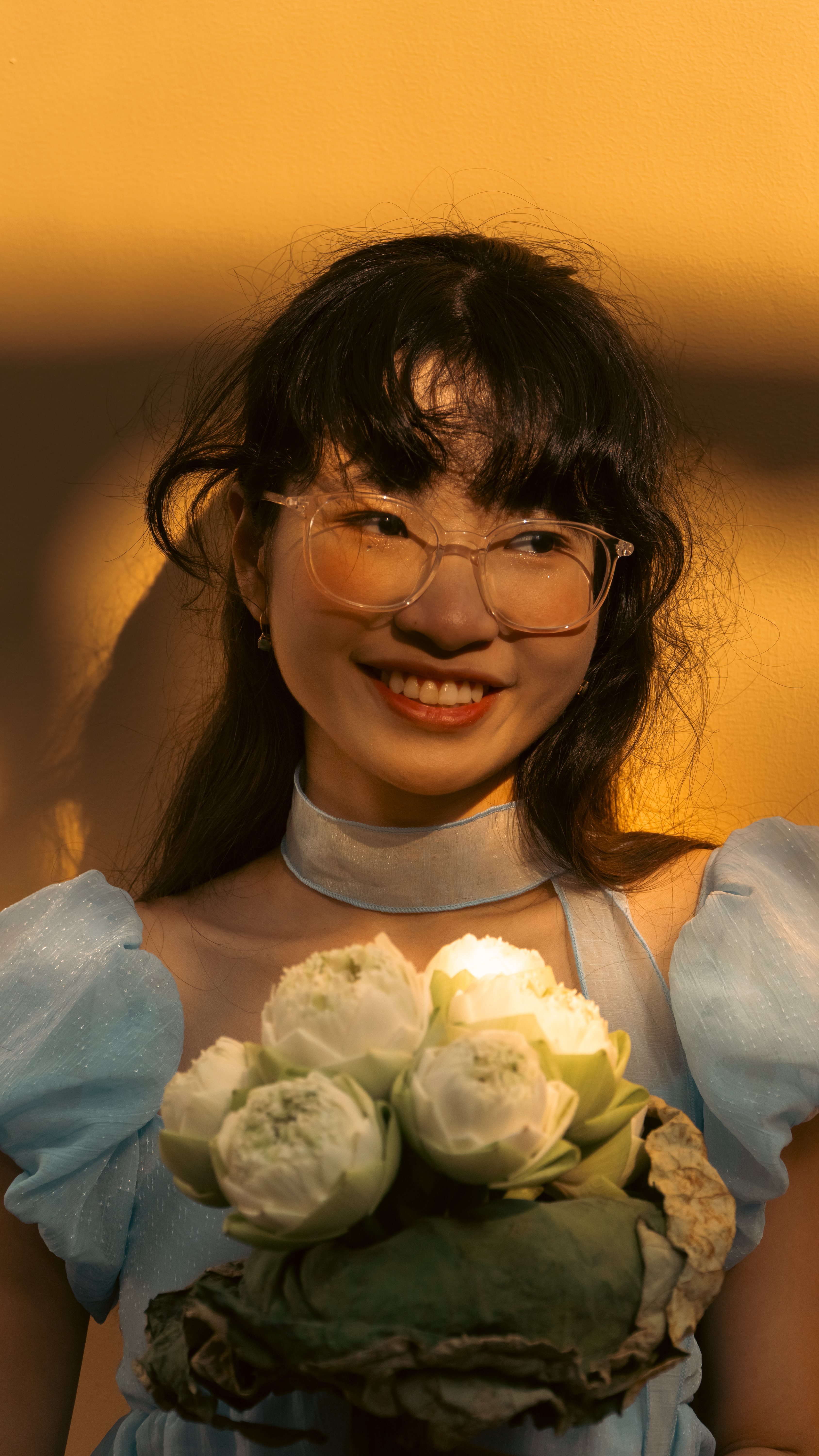 A woman wearing glasses and holding white flowers smiles warmly in soft lighting.