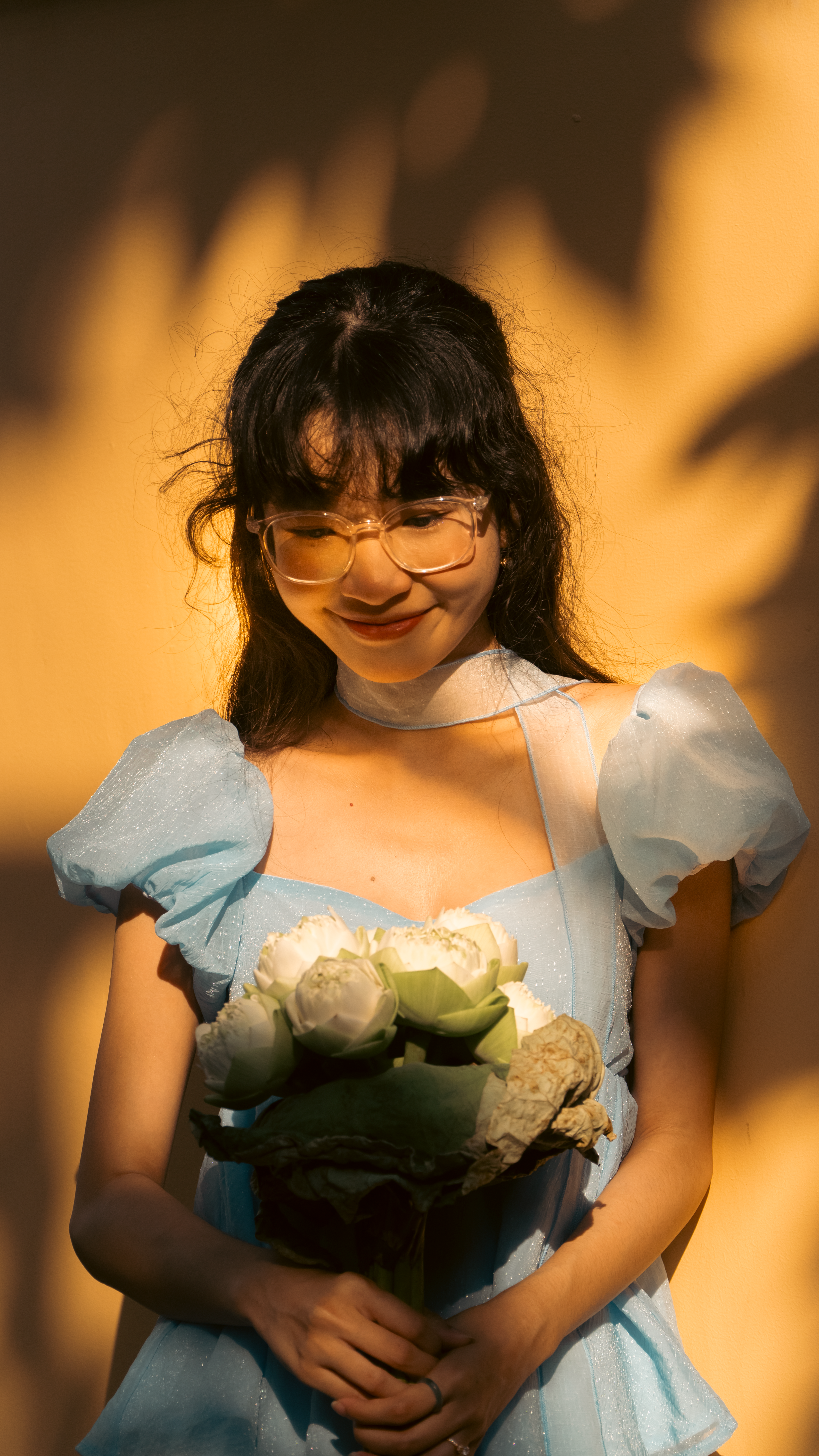 A young woman in a blue dress smiles while holding a bouquet of white flowers in warm light.