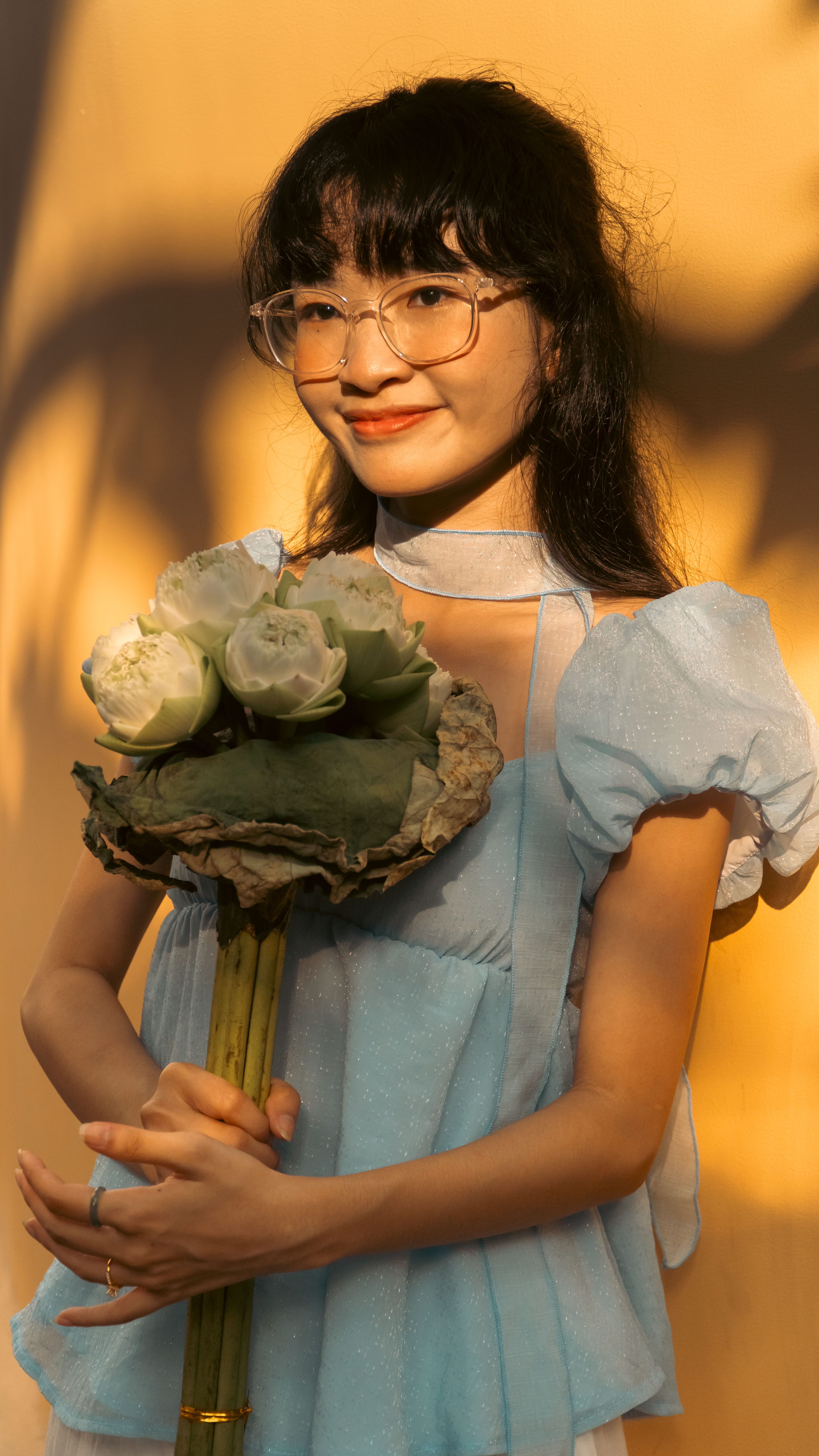 A young woman in a light blue dress holds lotus flowers, basking in warm sunlight.