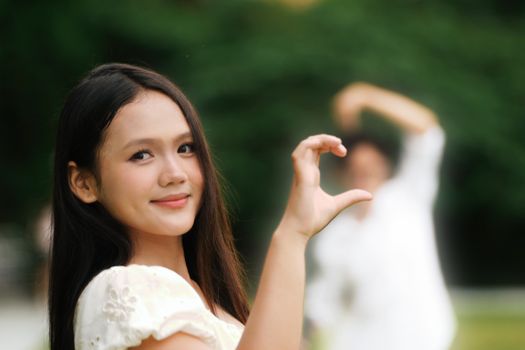 Young woman making a heart shape with hands, smiling outdoors. Blurred background shows another person.