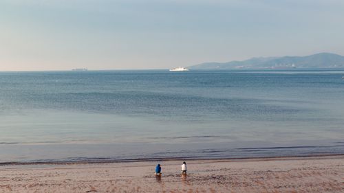 Free Serene beach scene with two people enjoying the view, distant ships, and a clear horizon. Stock Photo