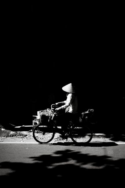 A cyclist with a conical hat rides through a shadowed street in Hà Nội, Vietnam.
