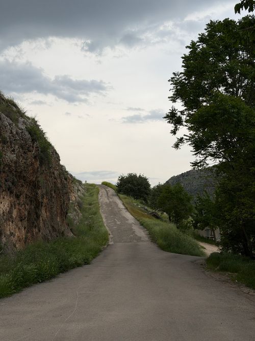 Free A peaceful country road surrounded by lush greenery and rocky hills on a cloudy day. Stock Photo