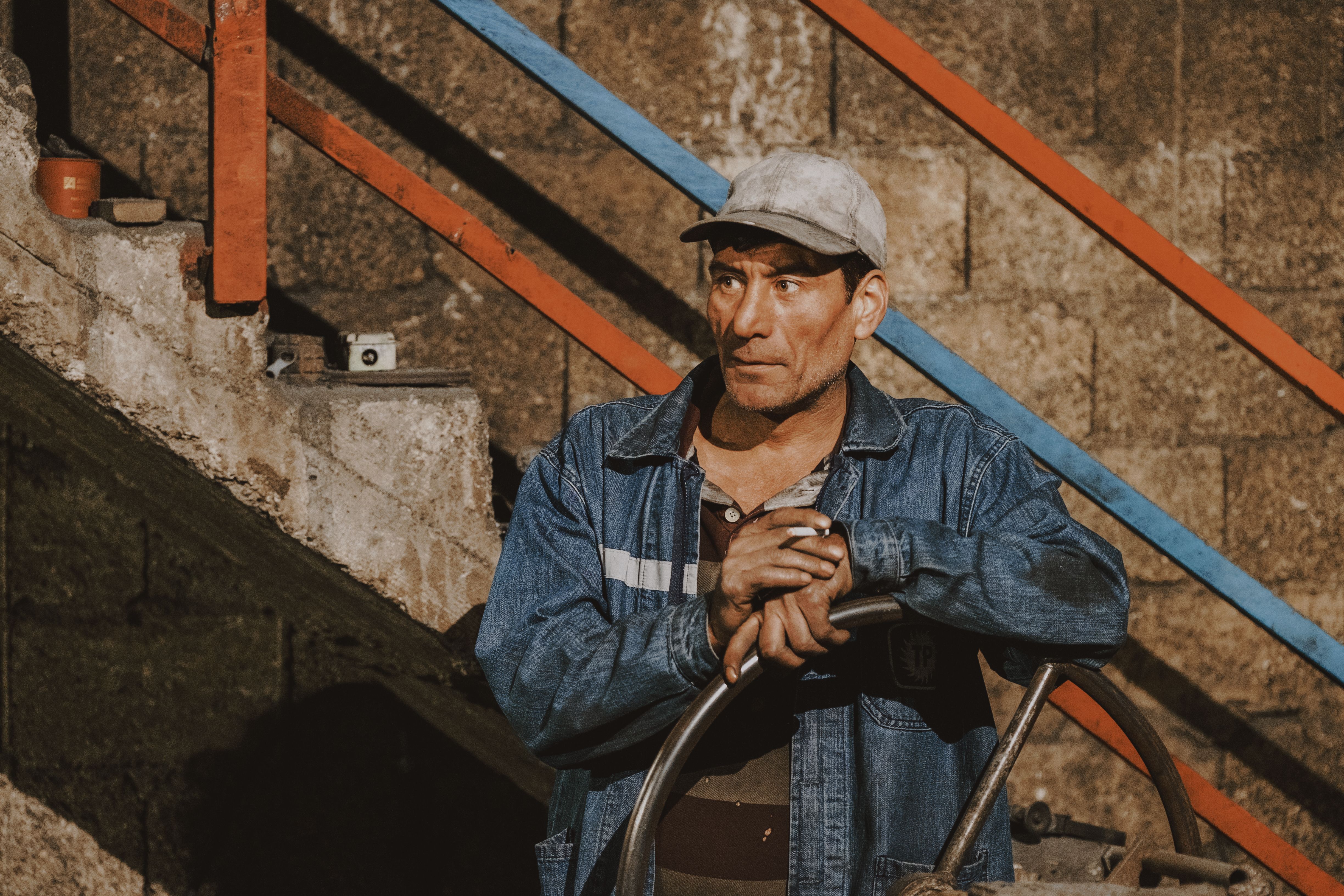 A rugged male worker in a denim jacket stands beside a colorful railing indoors.