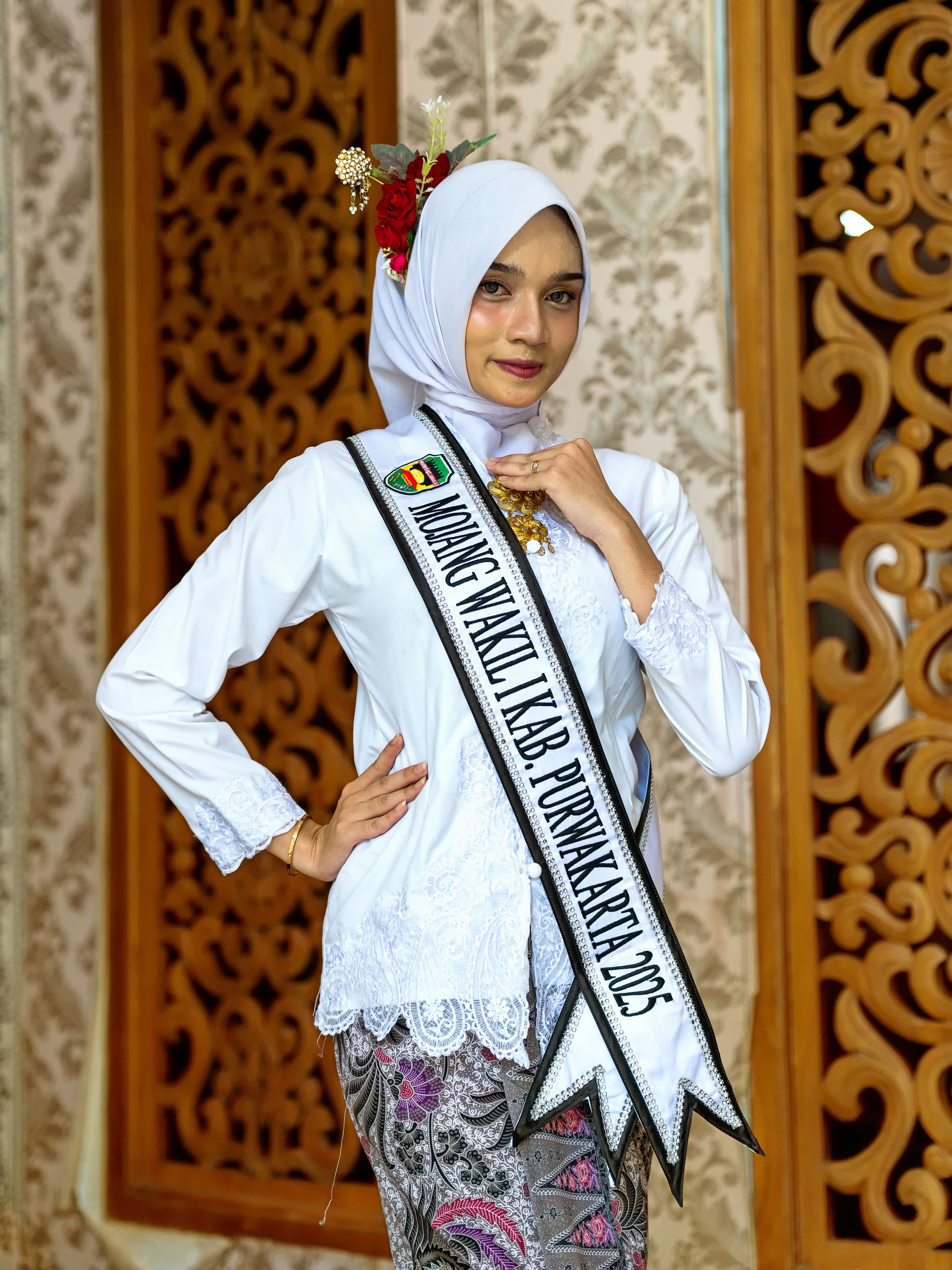 Woman in traditional dress and hijab with a sash posing elegantly indoors.