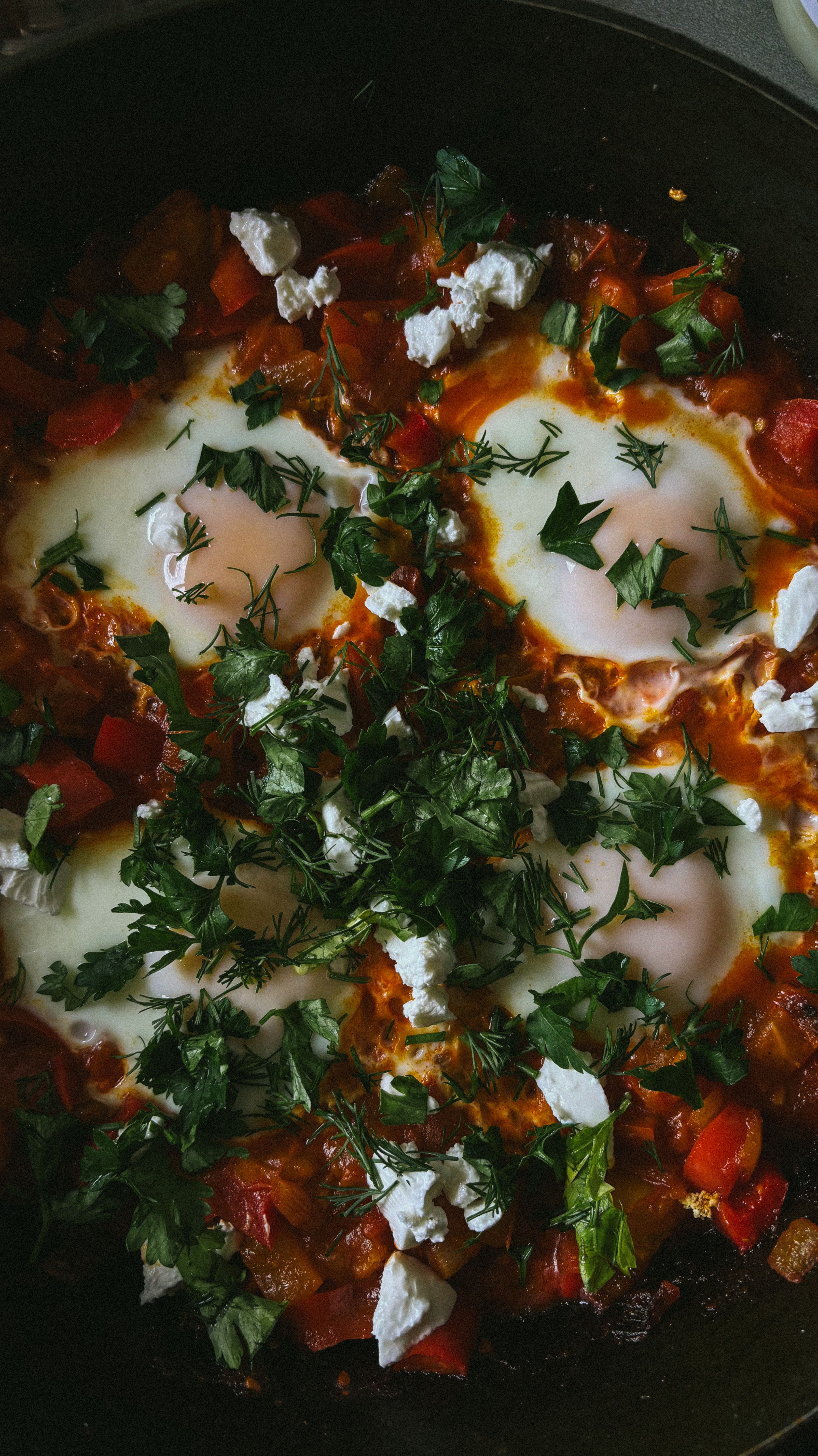 Six eggs nestled in bubbling red tomato sauce with visible green chile pieces, topped with crumbled white feta cheese in a cast iron skillet