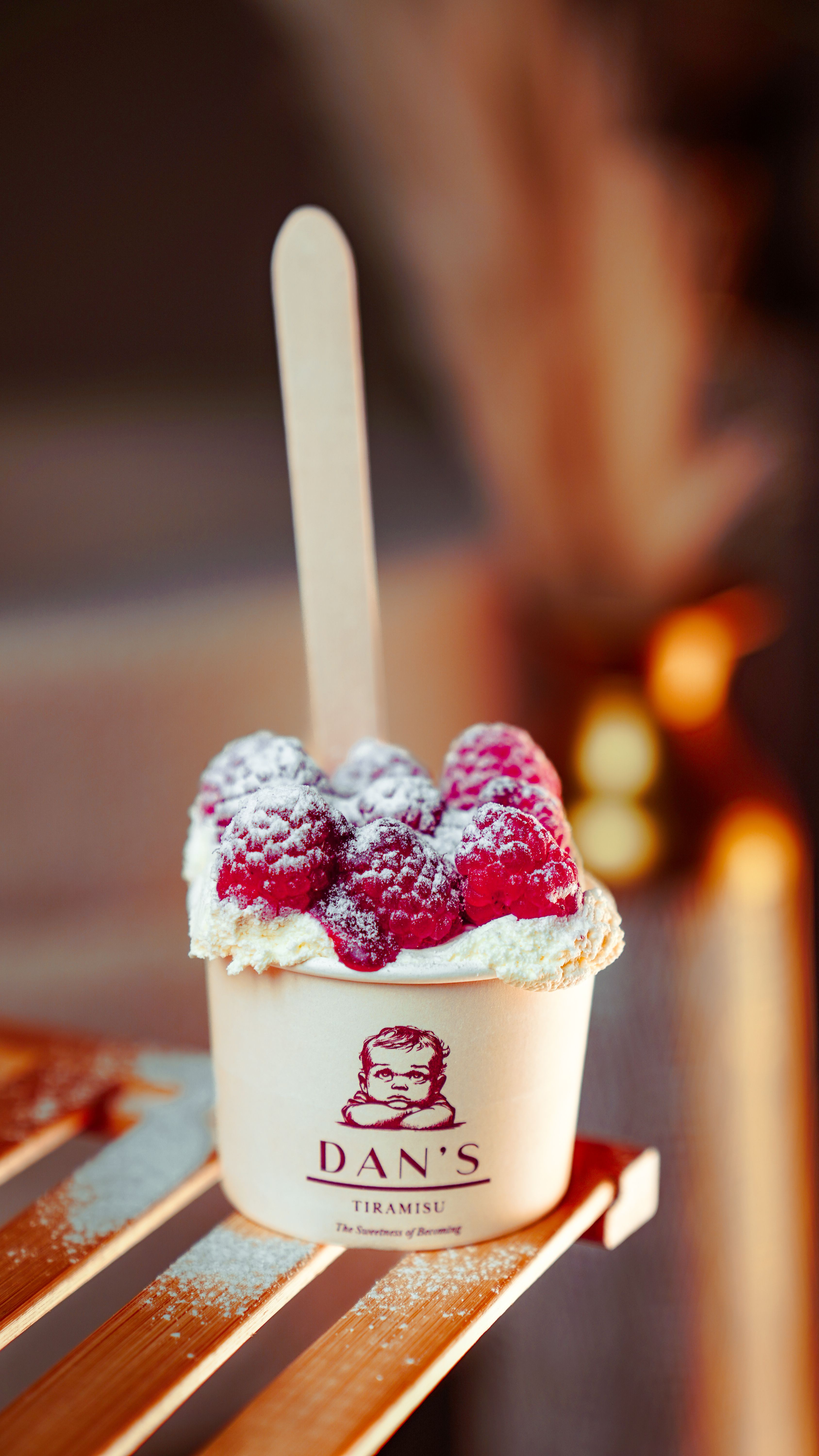 Tiramisu dessert topped with fresh raspberries in a branded cup with a wooden spoon.