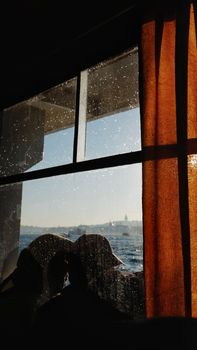 Silhouetted people viewed through a rainy window on a ferry with a sea background.