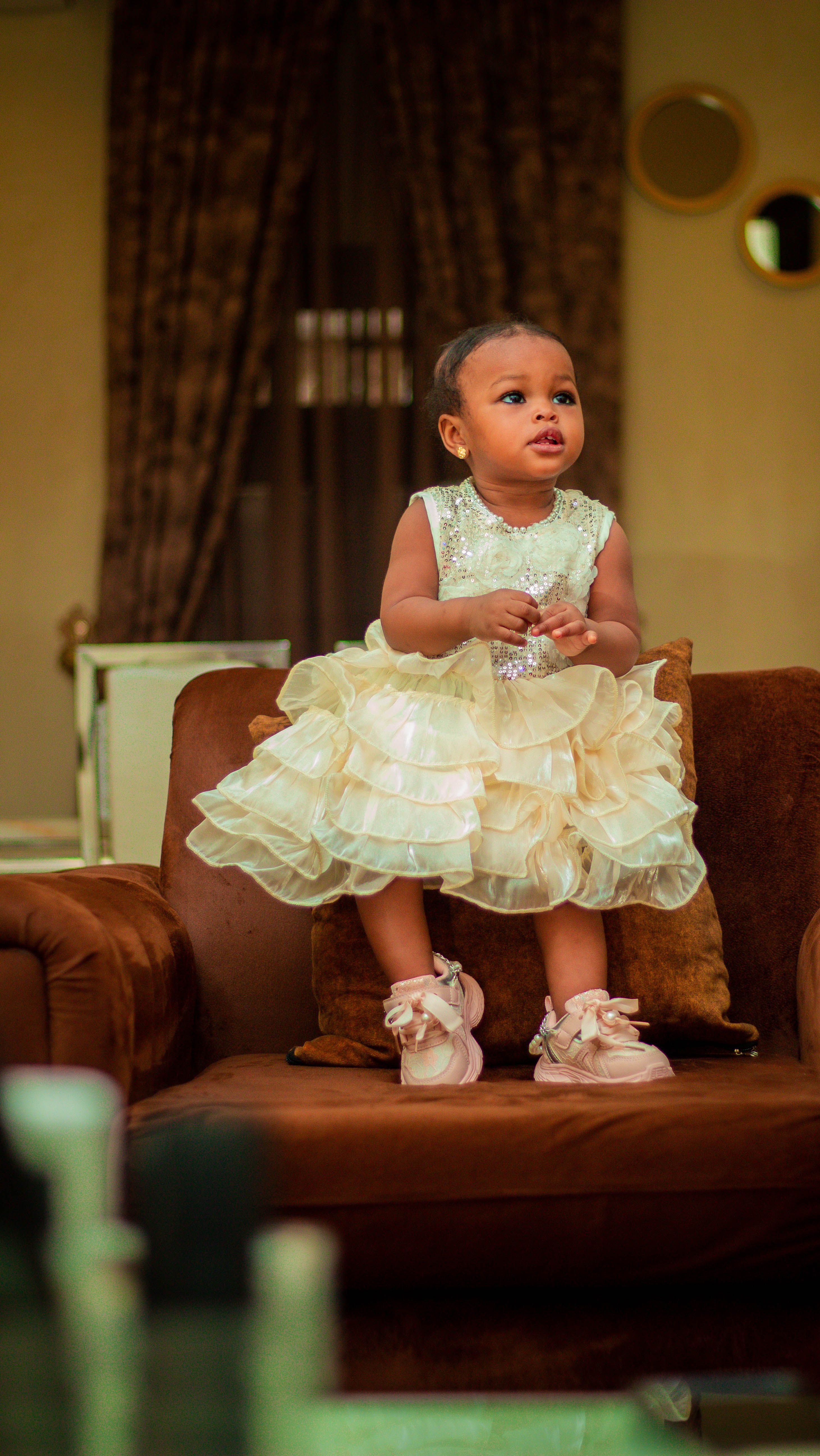 Cute baby girl in a fancy dress standing on a sofa indoors.