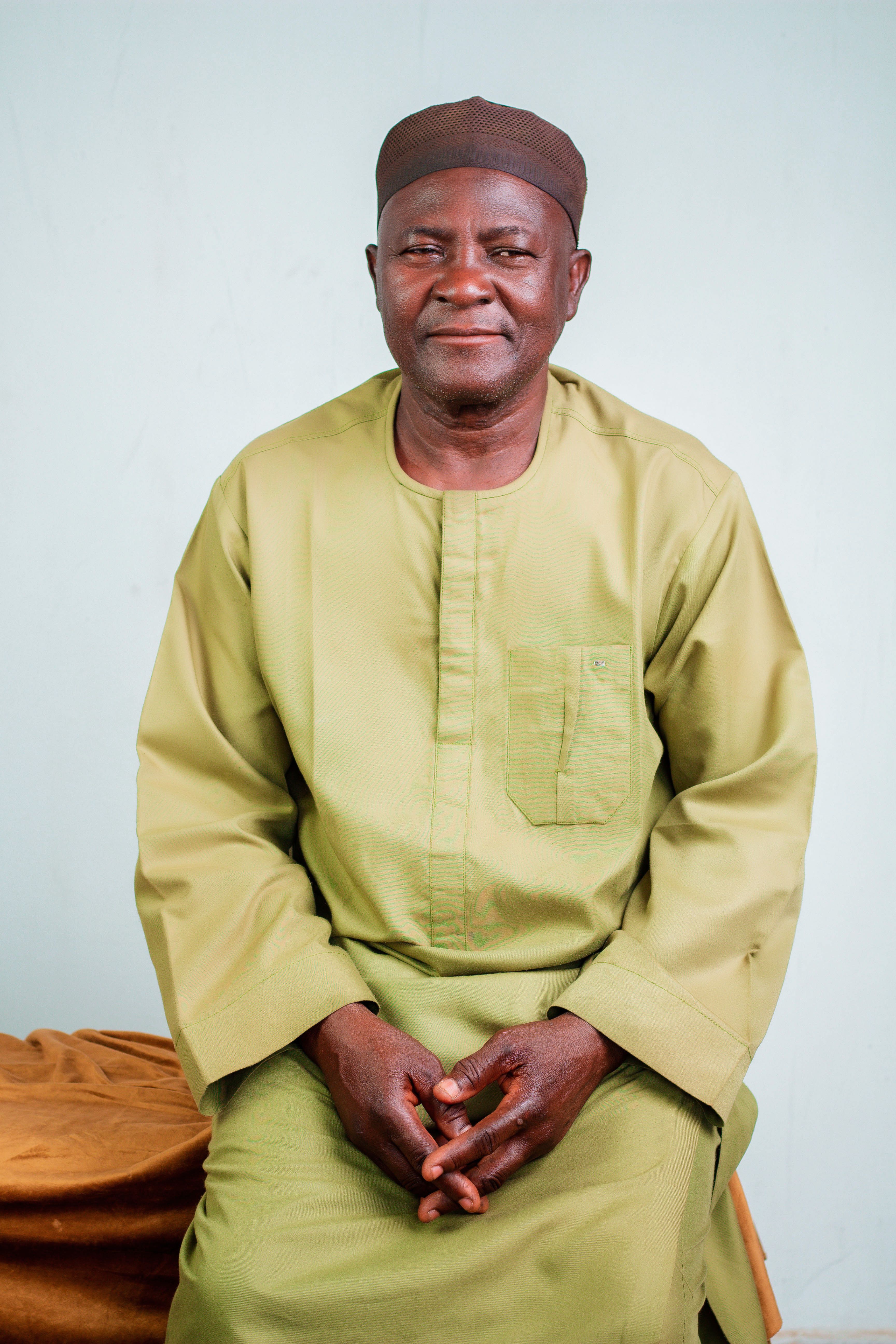 Elderly man wearing traditional green attire, seated indoors.