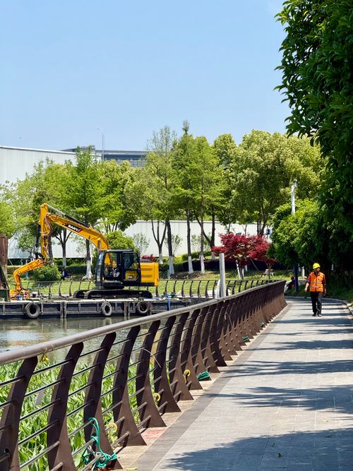 Free A construction worker walks in a park beside a digger under bright sunlight. Stock Photo