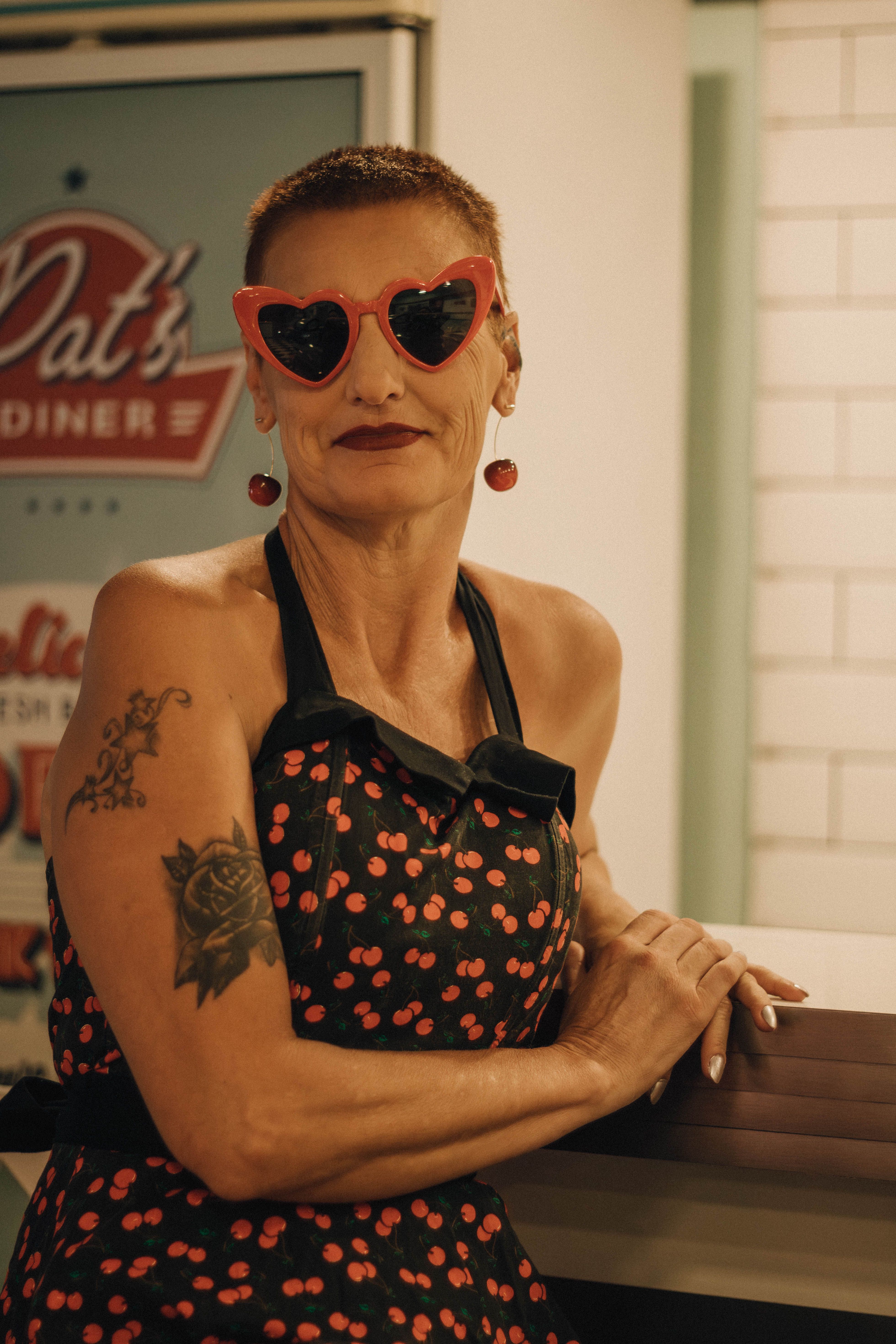 Stylish woman wearing heart-shaped sunglasses and cherry-patterned dress in vintage diner.