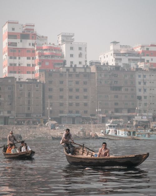 Free Men rowing wooden boats on a river in front of a densely populated urban area in Dhaka. Stock Photo