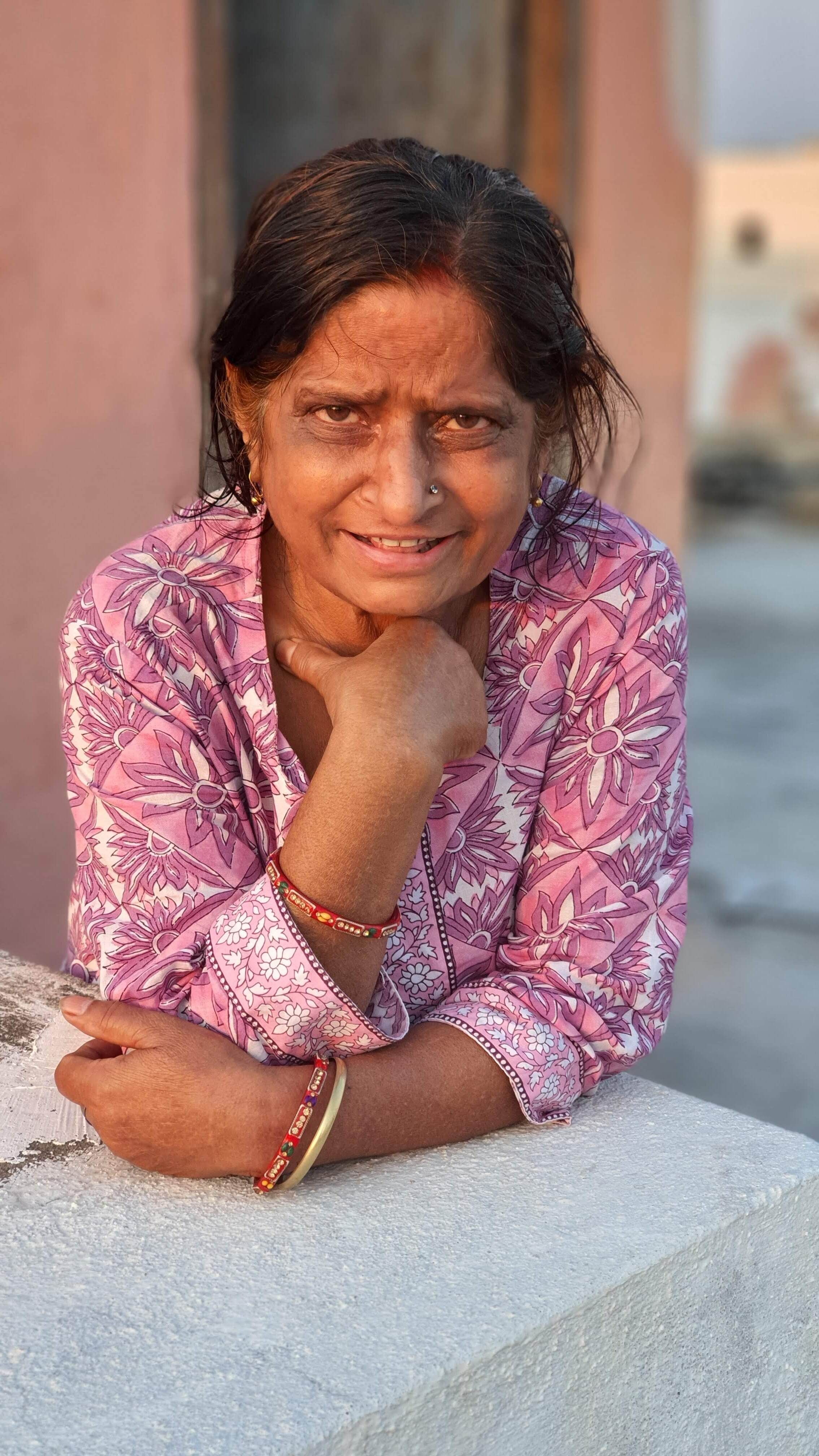 A senior woman leaning on a stone wall, smiling warmly in colorful attire.