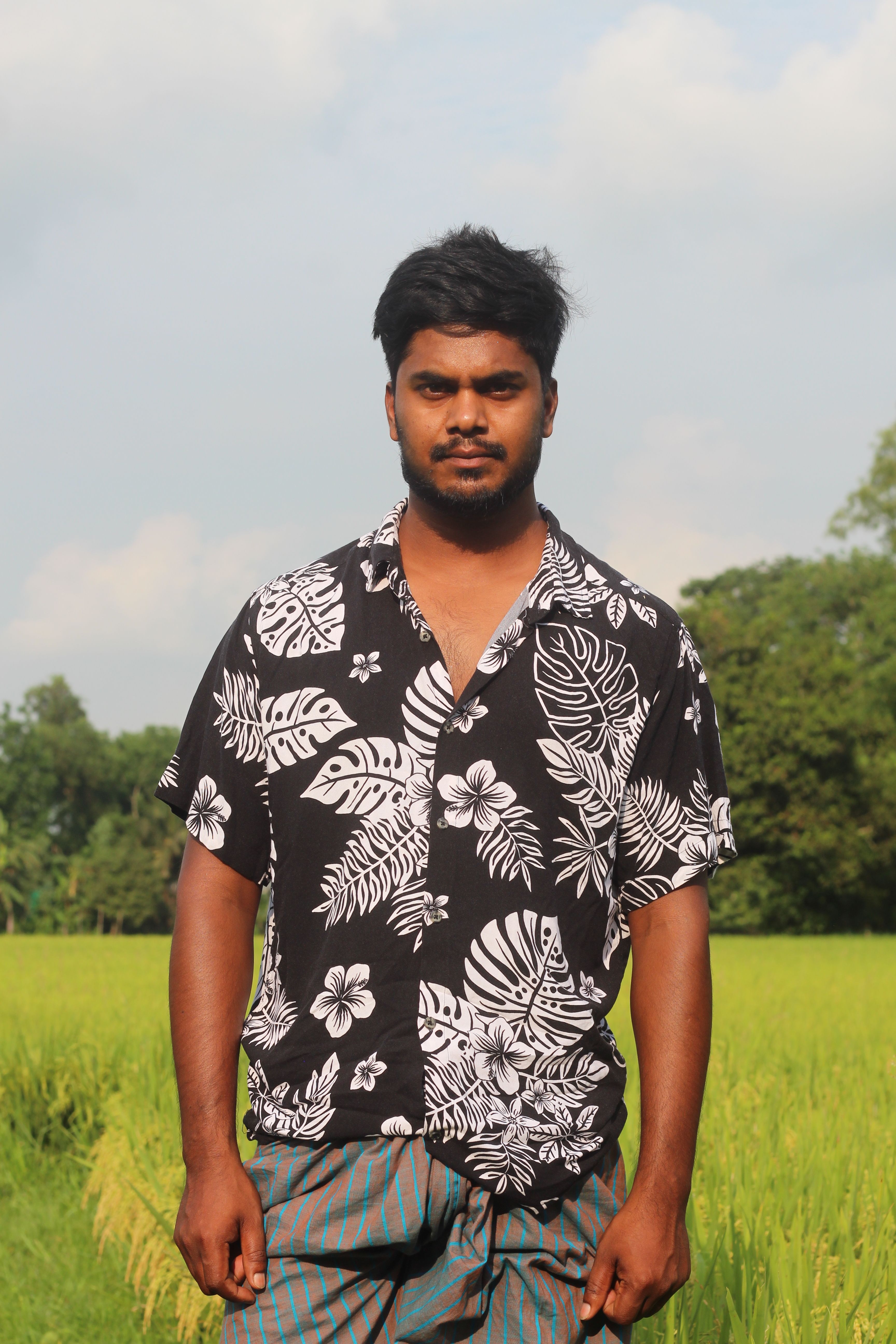 A man in a black and white floral shirt stands confidently in a sunny field.