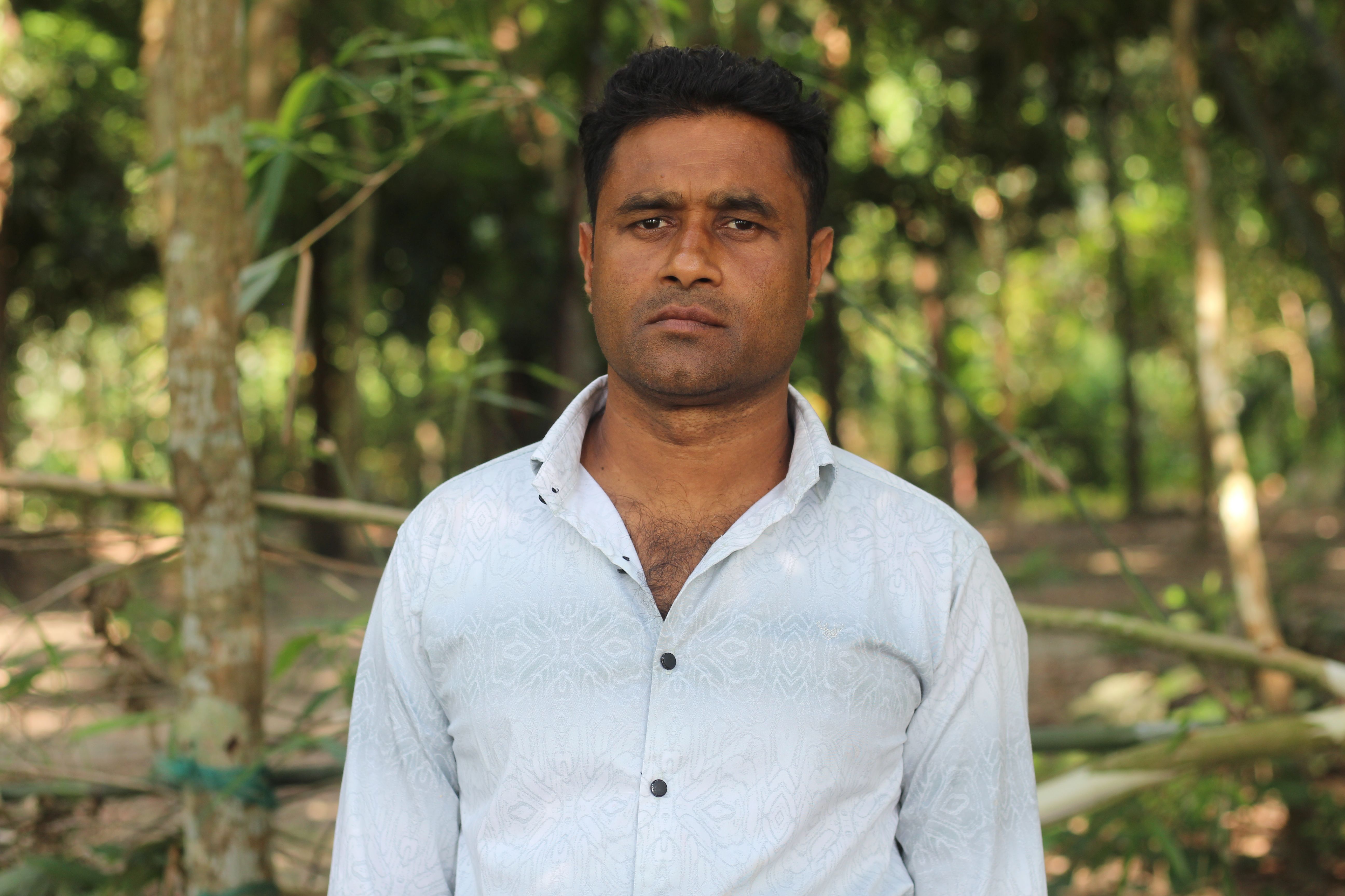 A confident man stands in a serene forest, wearing a white shirt, as sunlight filters through the trees.
