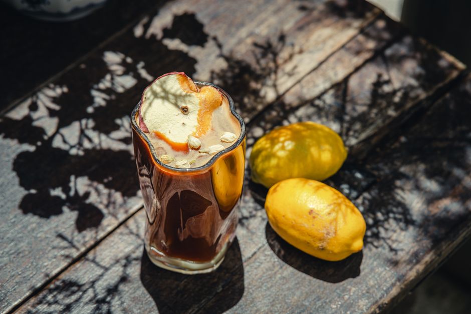A frothy iced coffee served with lemons on a rustic wooden table under natural light.