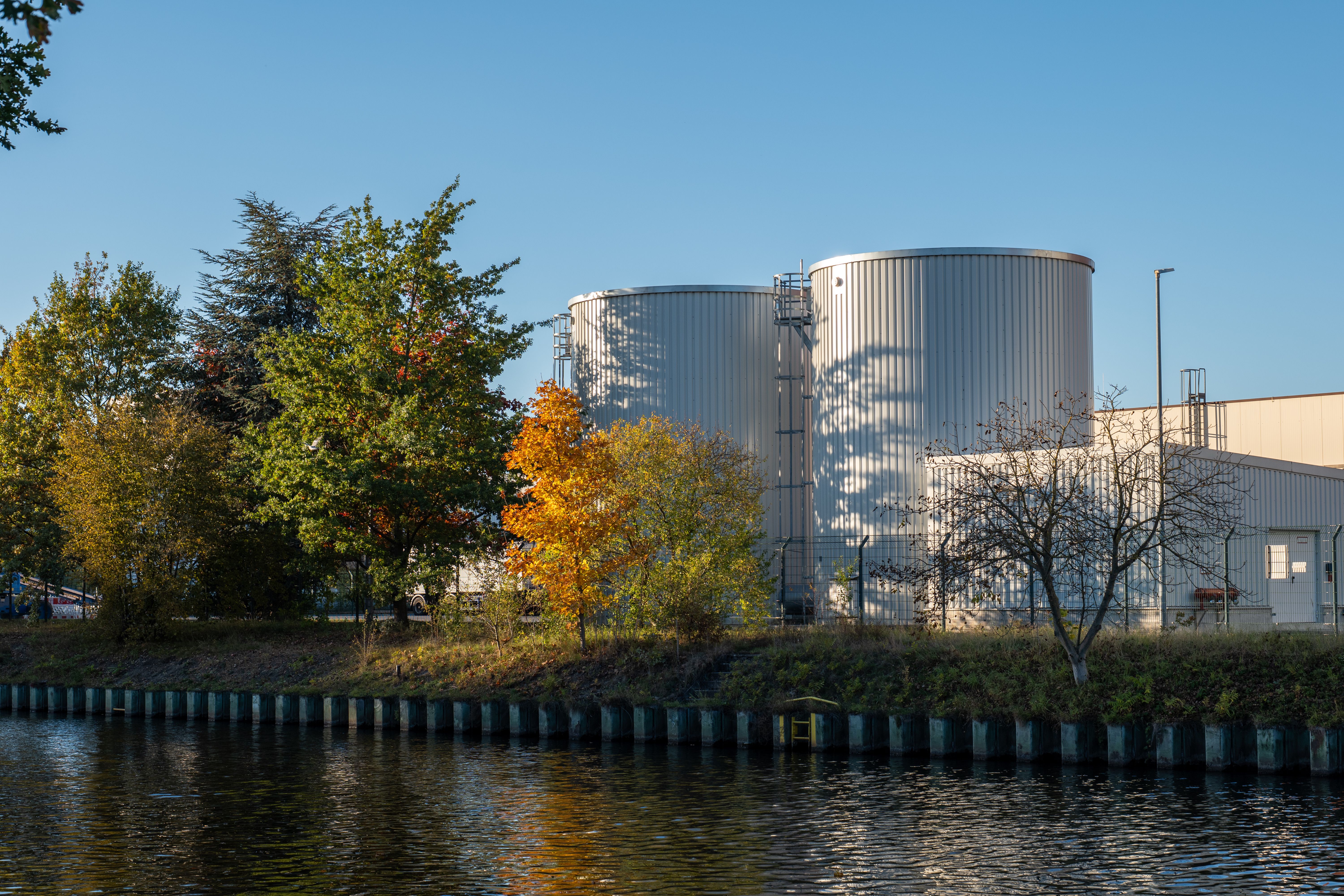 A scenic view of two large silos beside a tree-lined riverbank on a clear day.