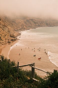 Surfers riding waves on a misty coastal beach with rugged cliffs during summer.