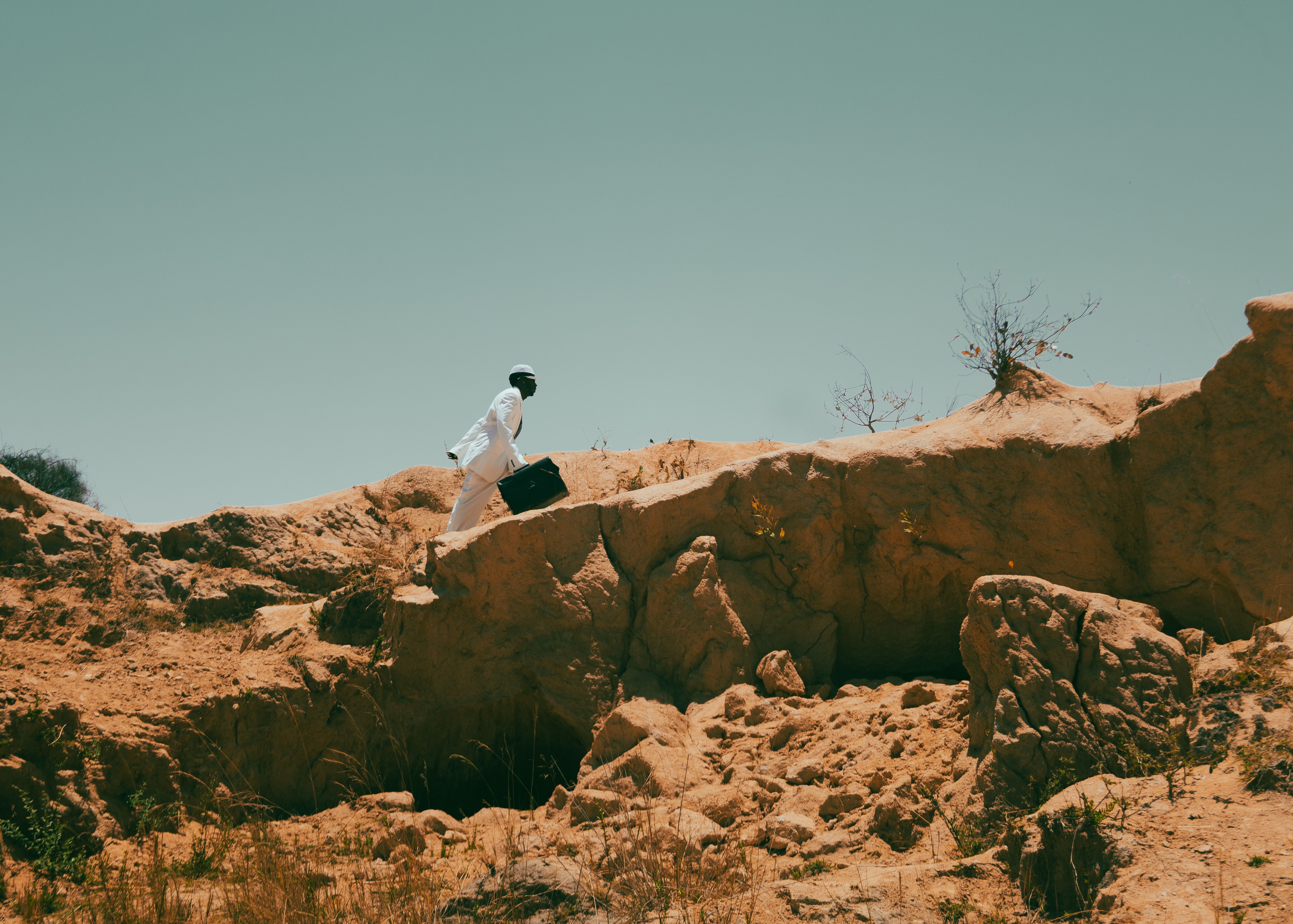 A man in white attire with a briefcase walking on a rocky, sunlit landscape.