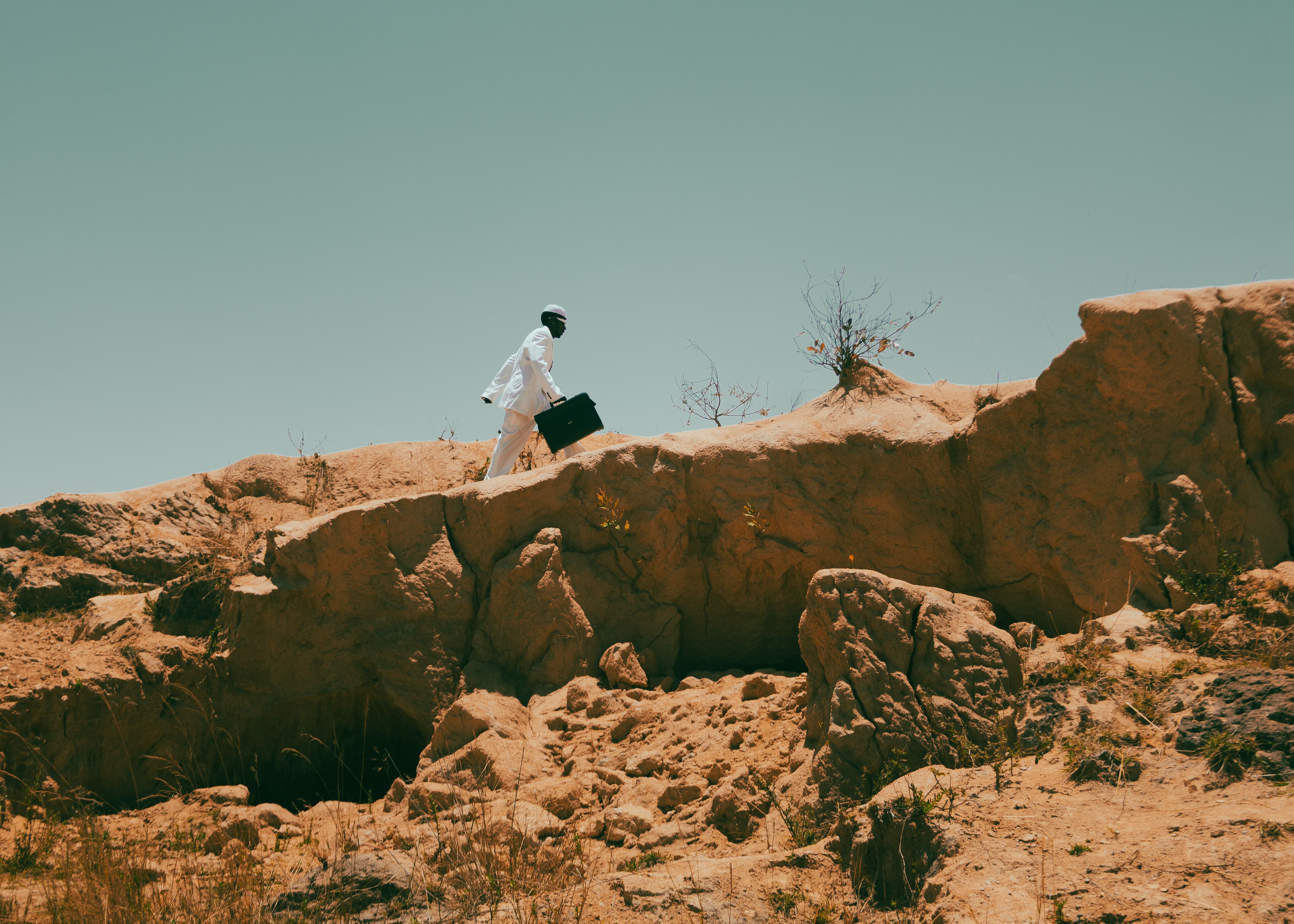 A man in white attire climbs a rugged landscape holding a briefcase, under a clear sky.