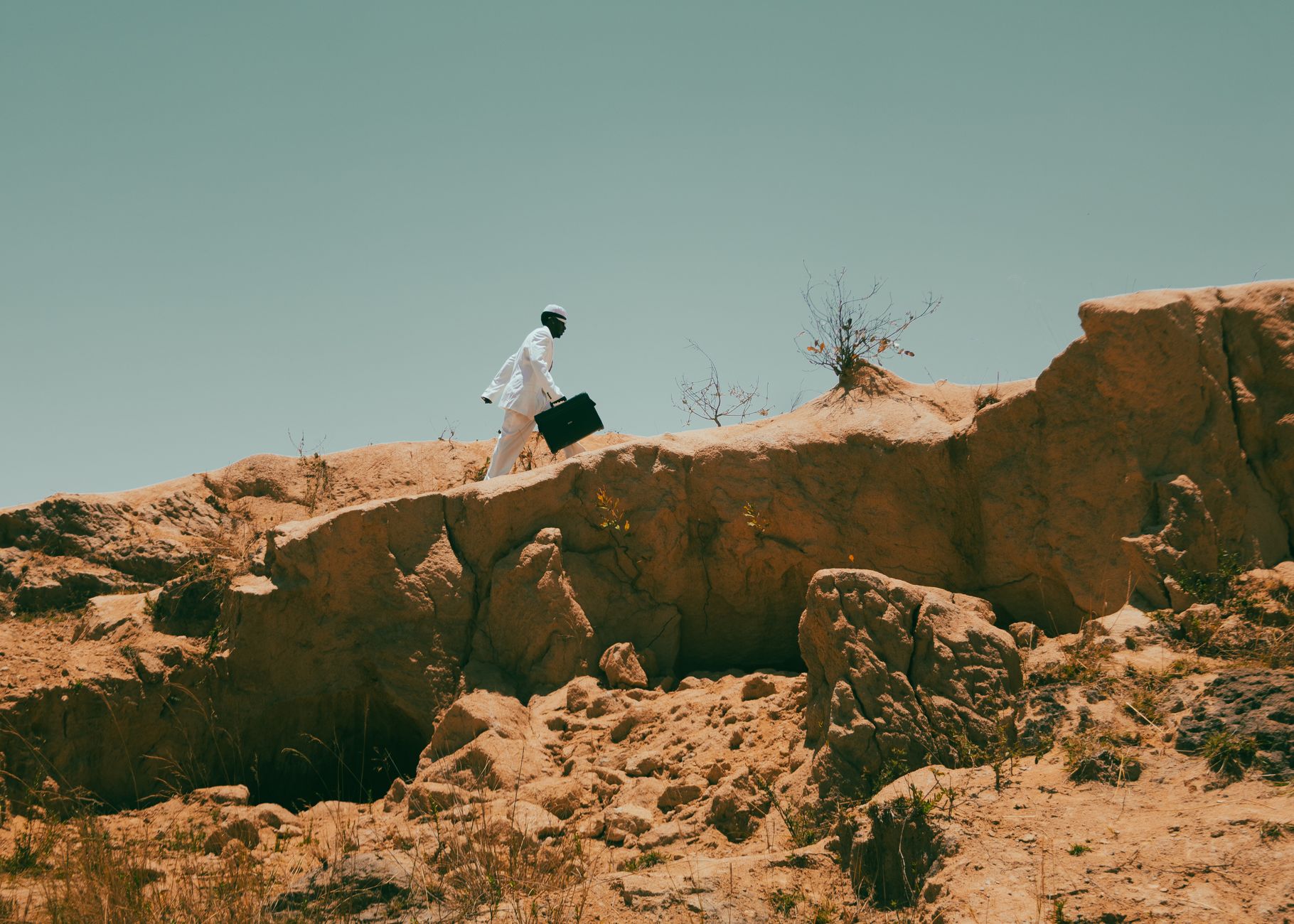 A man in white attire climbs a rugged landscape holding a briefcase, under a clear sky.