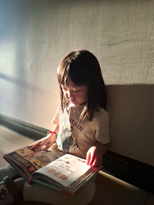 Free A young child reading a book while sitting against a sunlit wall indoors. Stock Photo