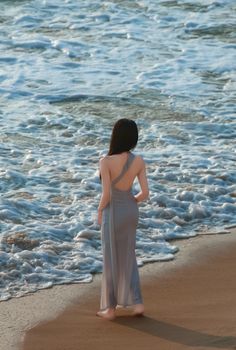 A woman in a flowing dress stands at the ocean's edge, contemplating the waves.