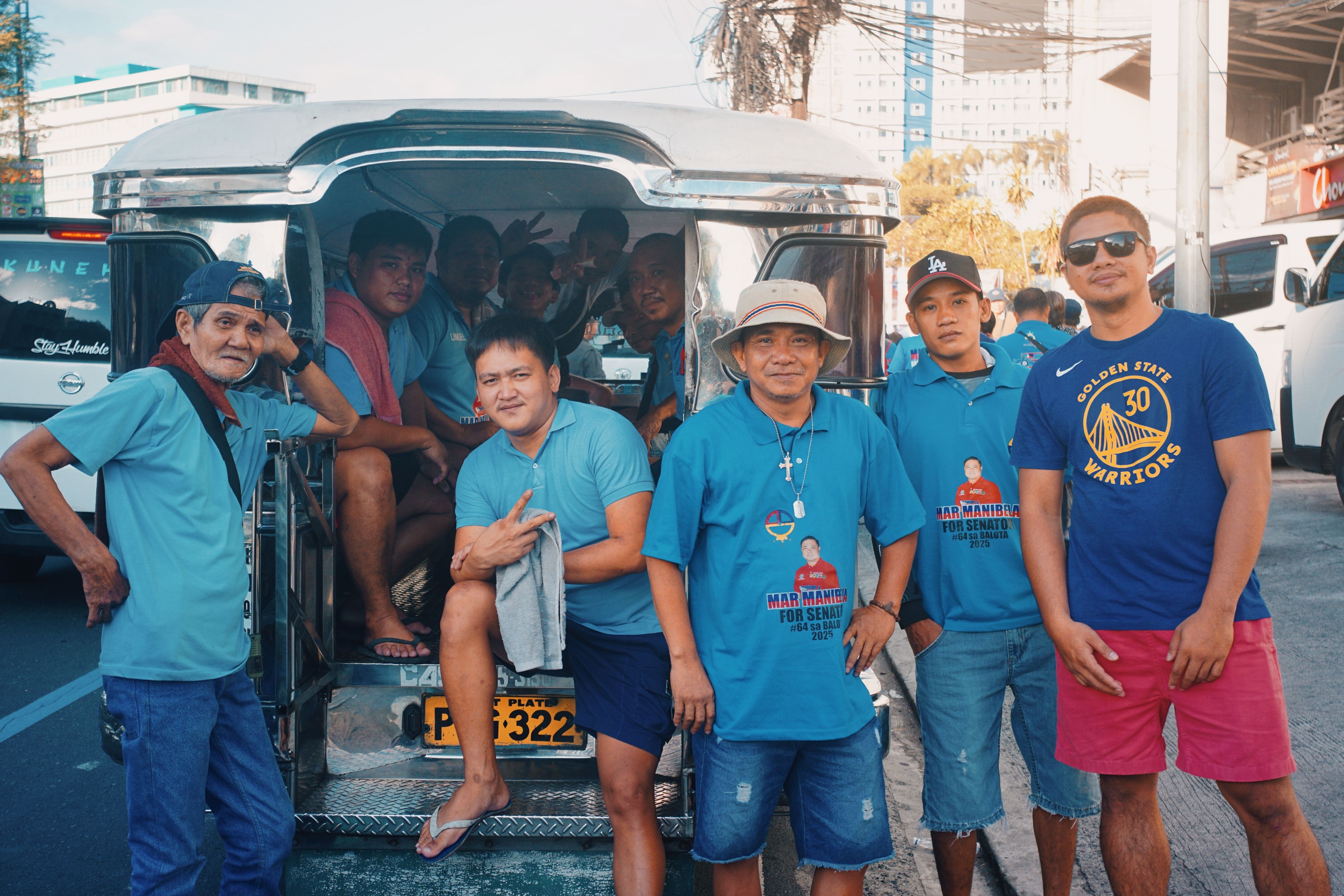 A group of men wearing blue shirts posing beside a jeepney on a city street during the day.