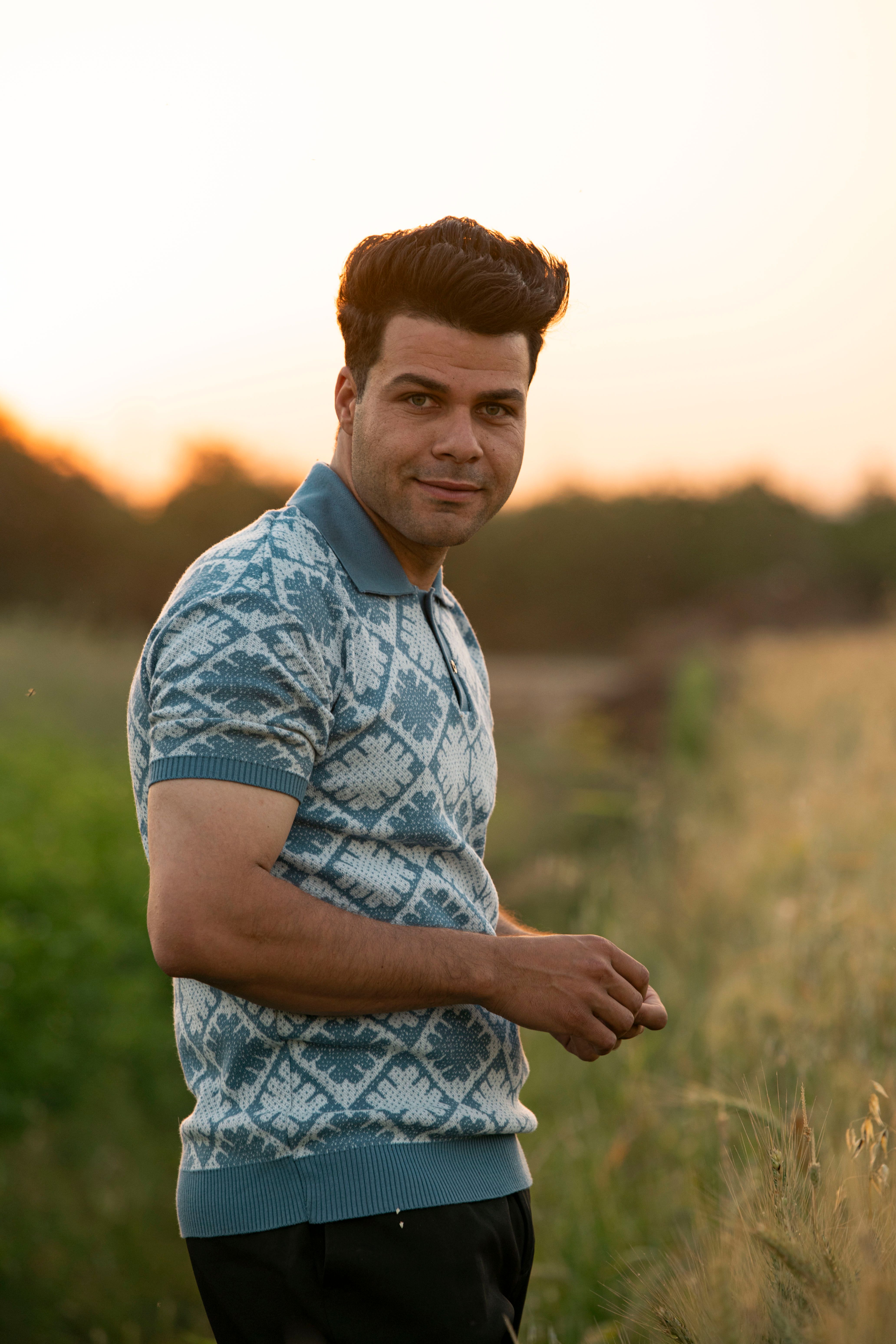 A man in a patterned shirt stands outdoors in a field at sunset.
