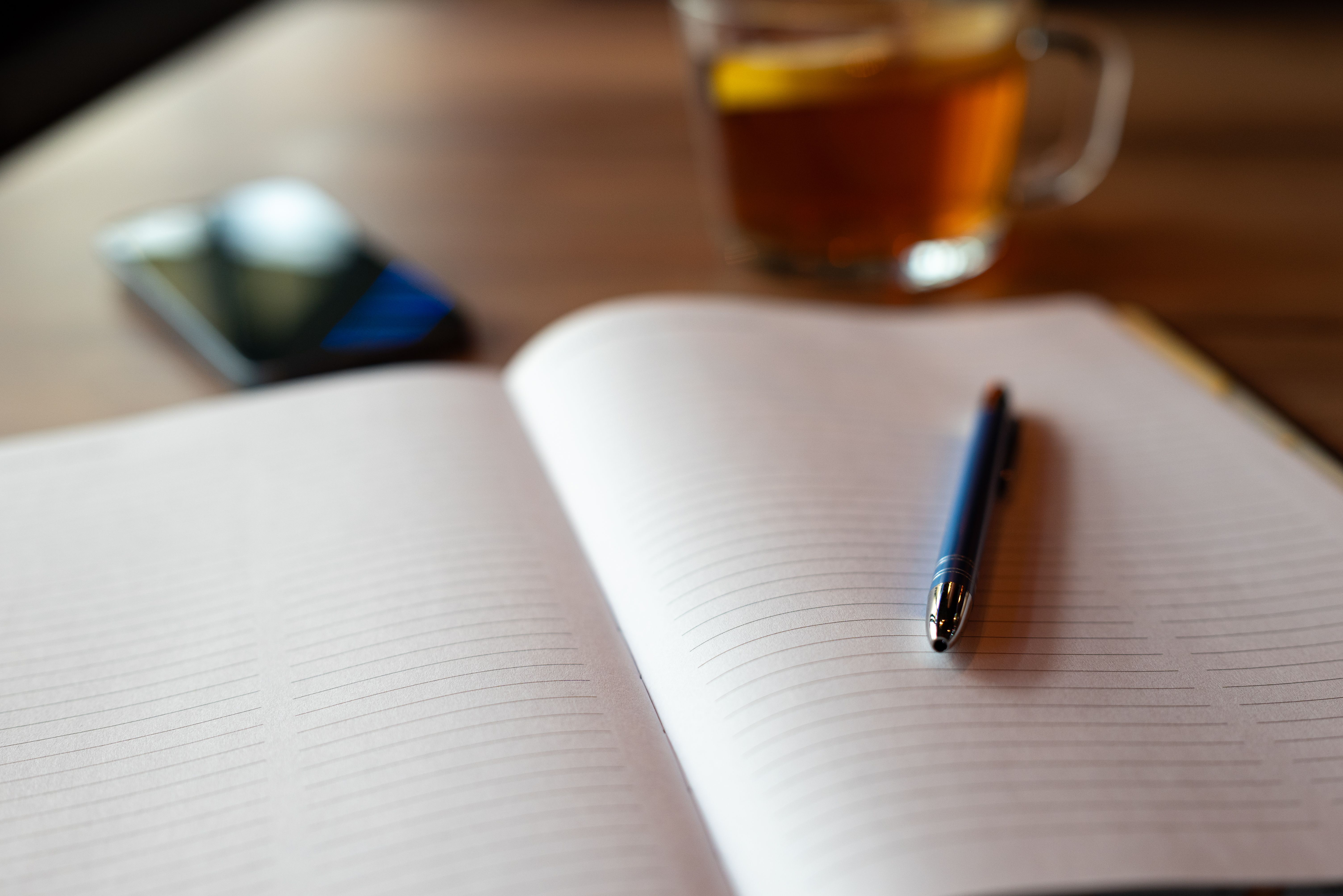 smartphone face down on a table next to a notebook and pen, with a cup of tea in background