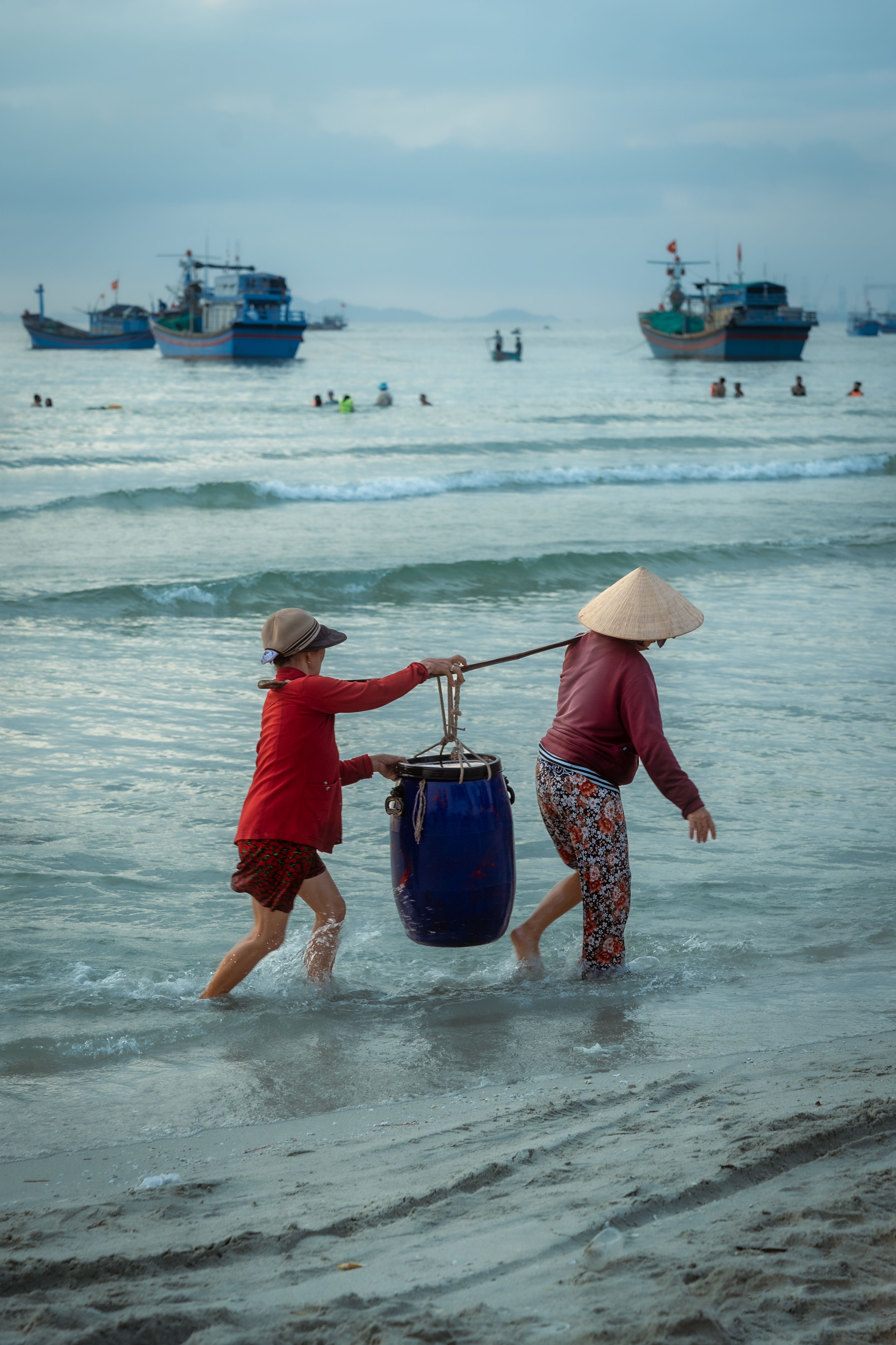 Two fishermen carrying a basket through shallow waters at Đông Ninh Hòa, Vietnam, with boats in the background.