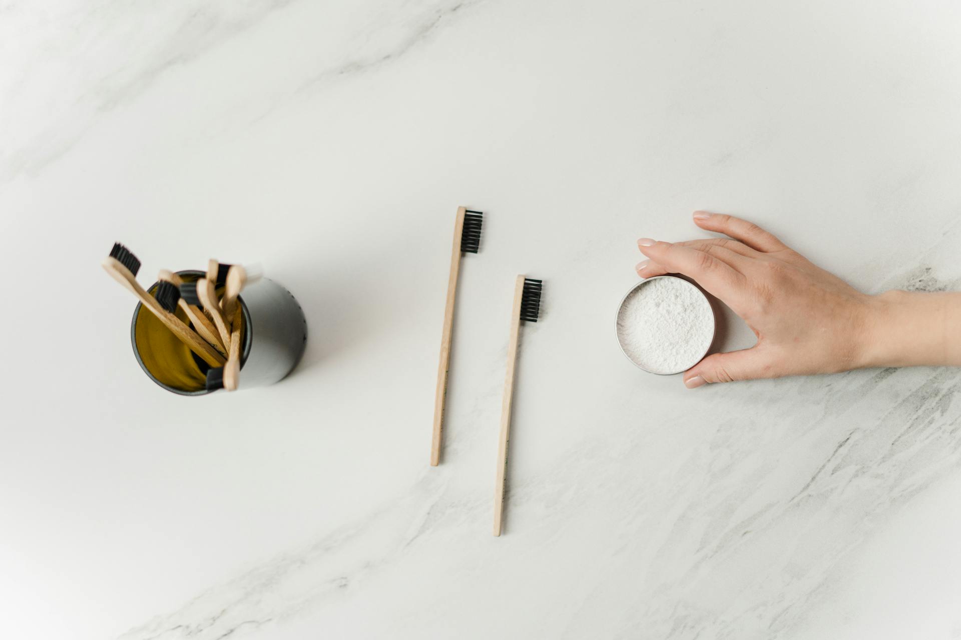 Person Holding Tooth Powder Jar Beside Bamboo Tooth Brushes