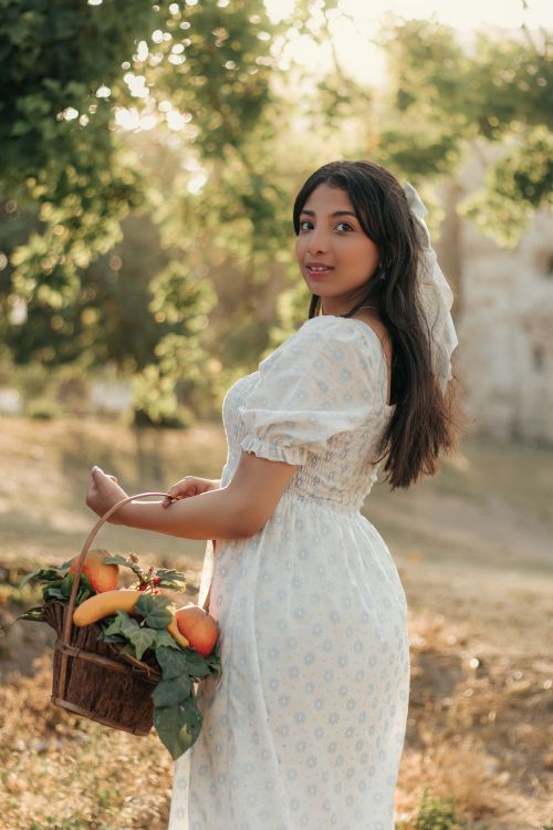Free Young woman in a vintage dress stands outdoors with a basket of fresh produce under sunlit trees. Stock Photo