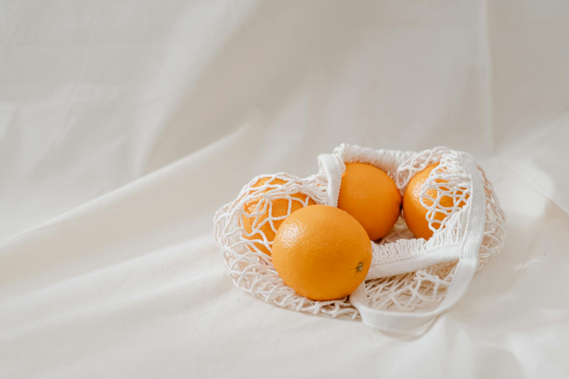 Fresh oranges placed in a sustainable mesh bag on white linen background.