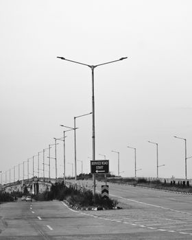 Black and white photo of an empty service road with streetlights and a sign at the start.