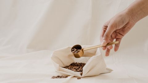 A hand scoops coffee beans from a cloth bag on soft white fabric, perfect for zero waste concept.