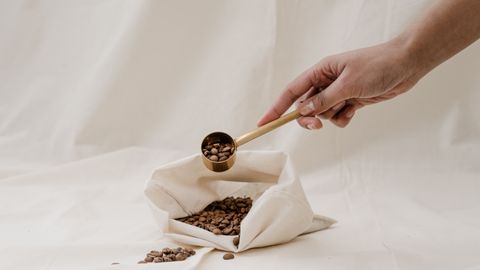 A hand holding a scoop over a fabric bag filled with coffee beans, highlighting a minimalist aesthetic.