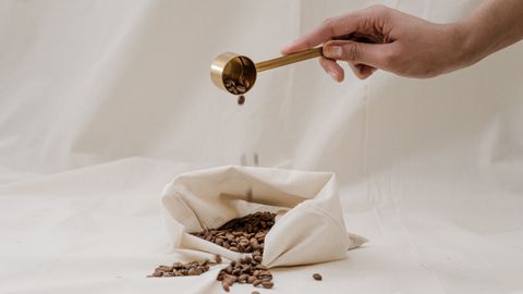 Hand pouring coffee beans with a gold scoop into an eco-friendly bag on a soft background.