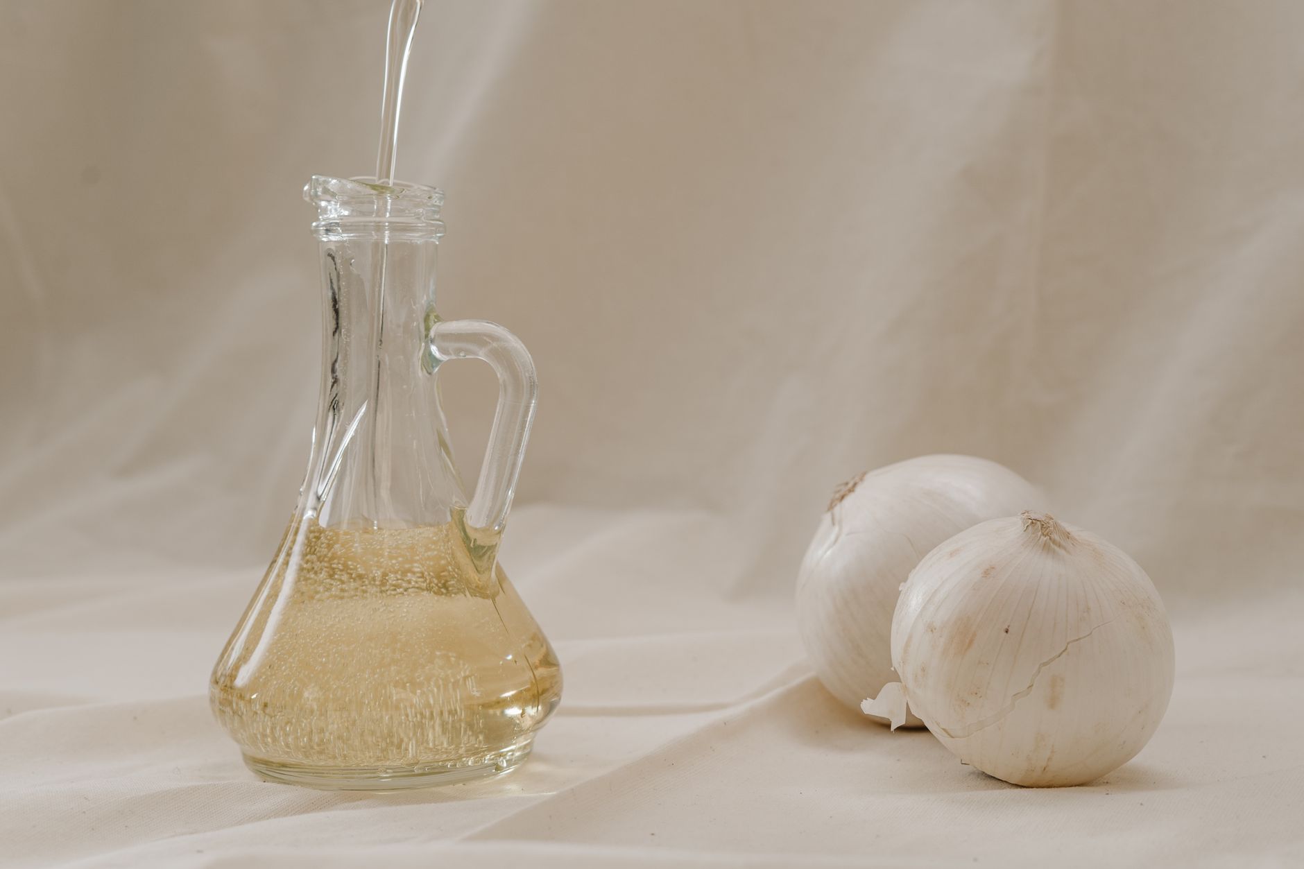 Close-up of a glass jar being filled with oil, alongside two garlic bulbs on a neutral background.