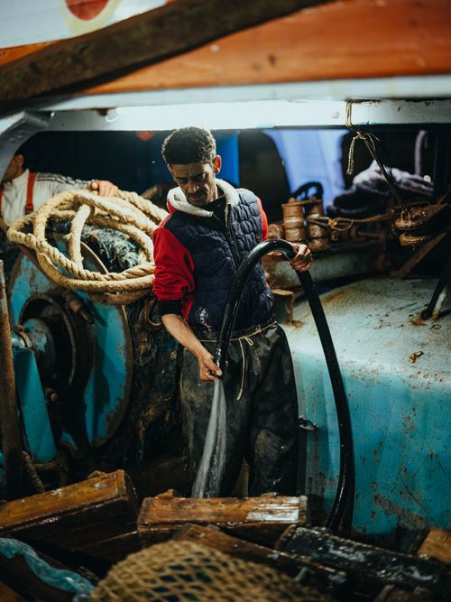 Free A fisherman cleans a boat deck with a hose under dim lighting, creating a rustic maritime scene. Stock Photo